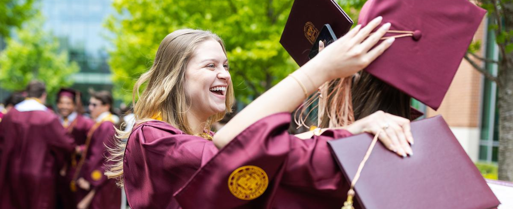 Two graduates in maroon caps and gowns embrace during an outdoor commencement